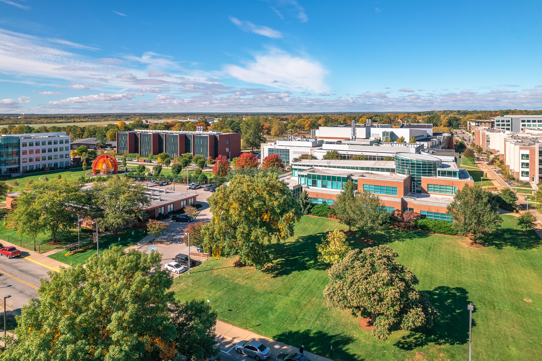 A large grassy area in the forefront gives way to 4 buildings in the district. Behind, trees spread out as far as the eye can see. - Discovery Park District - Purdue Research Foundation