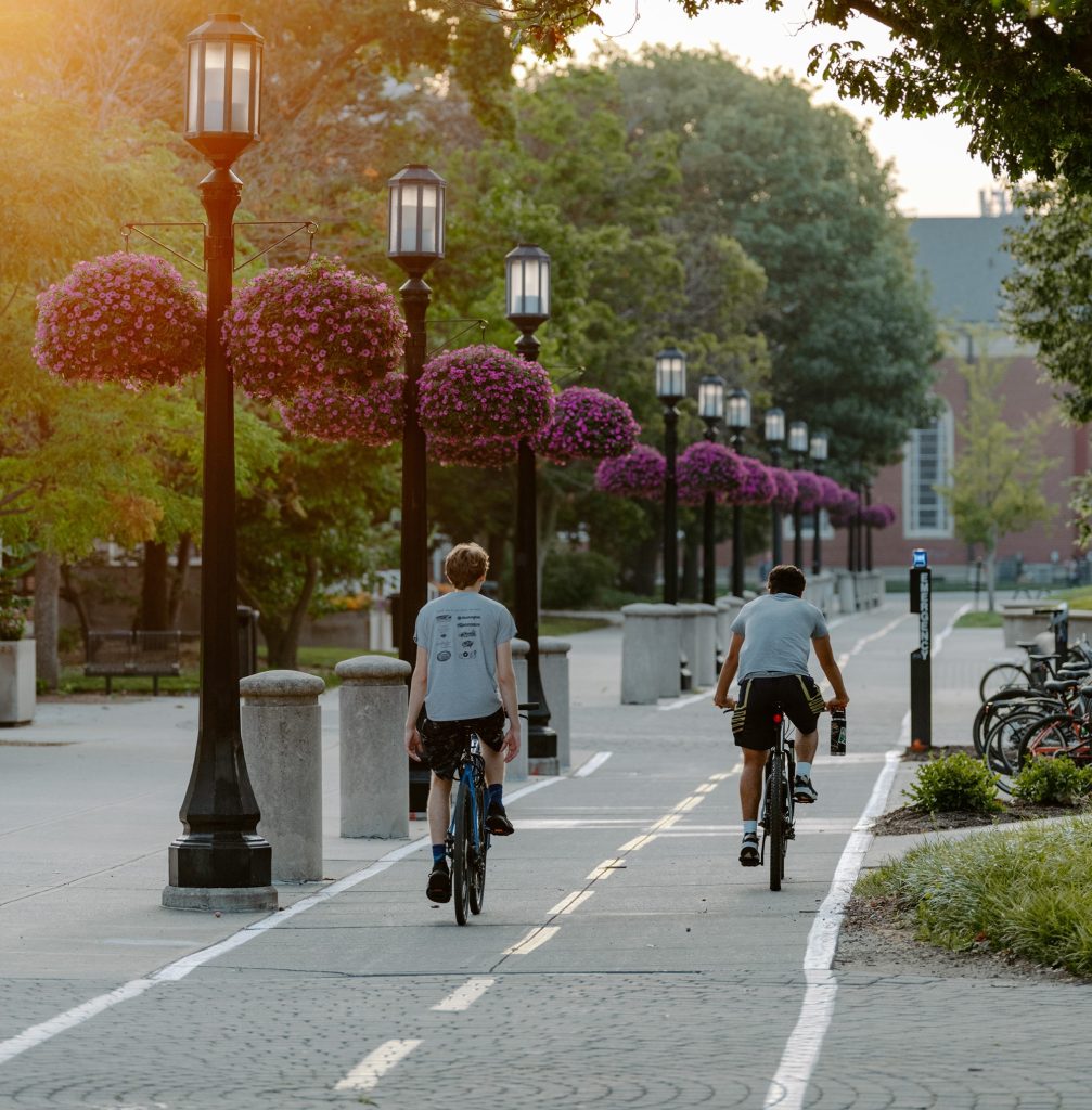 Two young people riding bikes away from the camera on a lamppost-lined bike trail on Purdue campus - Discovery Park District - Purdue Research Foundation