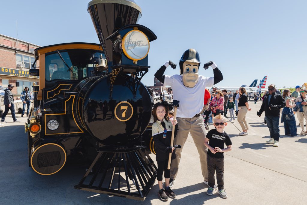 Purdue Pete stands between two small children next to the Boilermaker Special train at the Purdue University Airport - Discovery Park District - Purdue Research Foundation