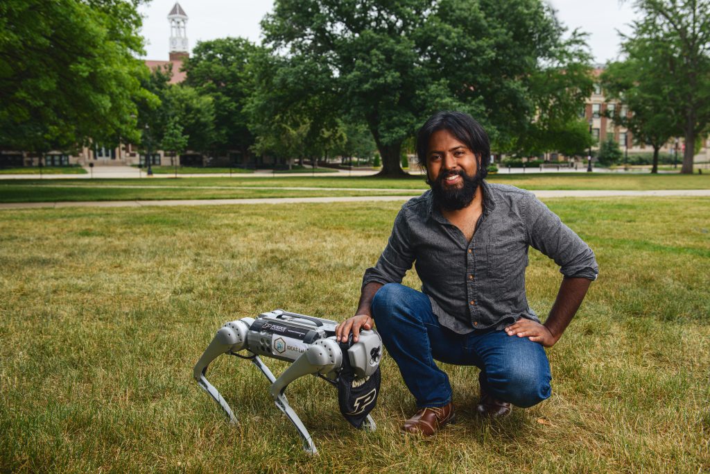 A man crouches next to a four-legged robot with a Purdue collar in a grassy field on Purdue's campus - Discovery Park District - Purdue Research Foundation
