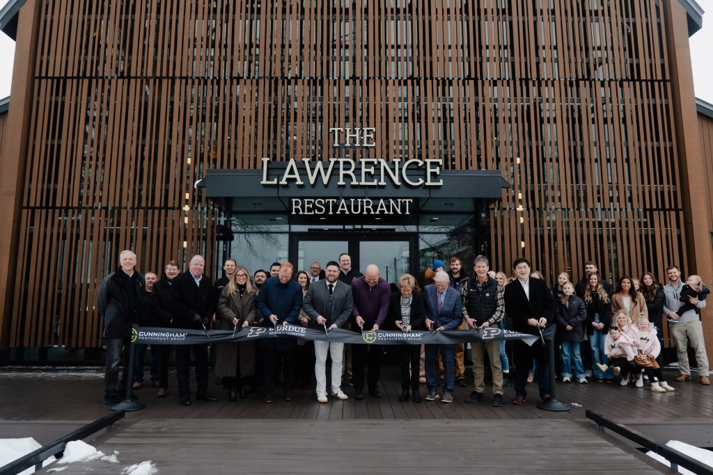 A large group of people stand, mid-ribbon-cutting, in front of the large wood and glass facade of The Lawrence Restaurant.