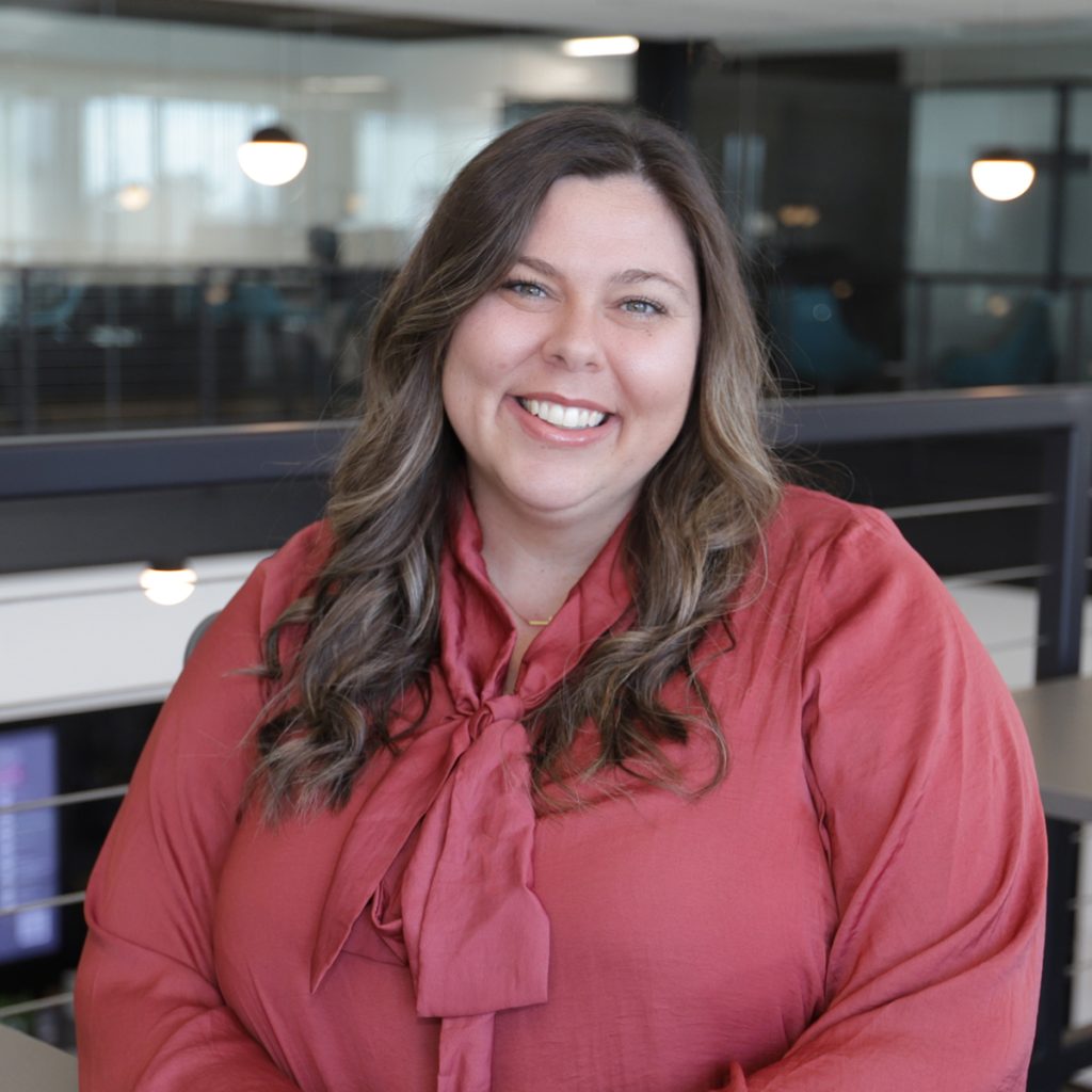 Smiling woman with wavy brown hair in a red blouse standing in a modern office setting.