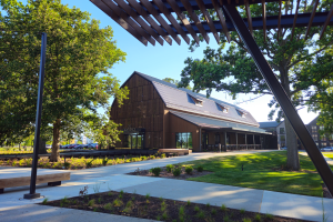 Modern barn-style building with glass facade, set amid lush green trees and manicured lawns under a clear blue sky.