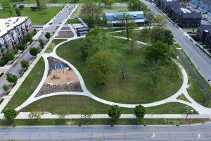 Aerial view of a green park with winding paths and scattered trees, surrounded by roads and modern apartment buildings, conveying a calm urban oasis.