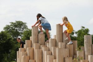 Two children climb wooden logs in a park under a cloudy sky with green trees in the background.