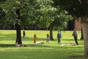 A group of people, including children and adults, play cornhole in a sunny park.