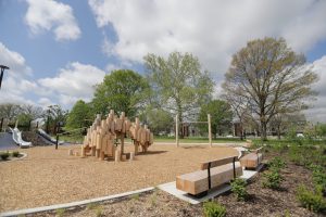 A playground with wooden climbing structures sits on mulch, surrounded by greenery. Benches line the area under a partly cloudy sky, conveying a peaceful atmosphere.