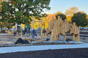 Two construction workers stand next to a wooden playground structure on a construction site.